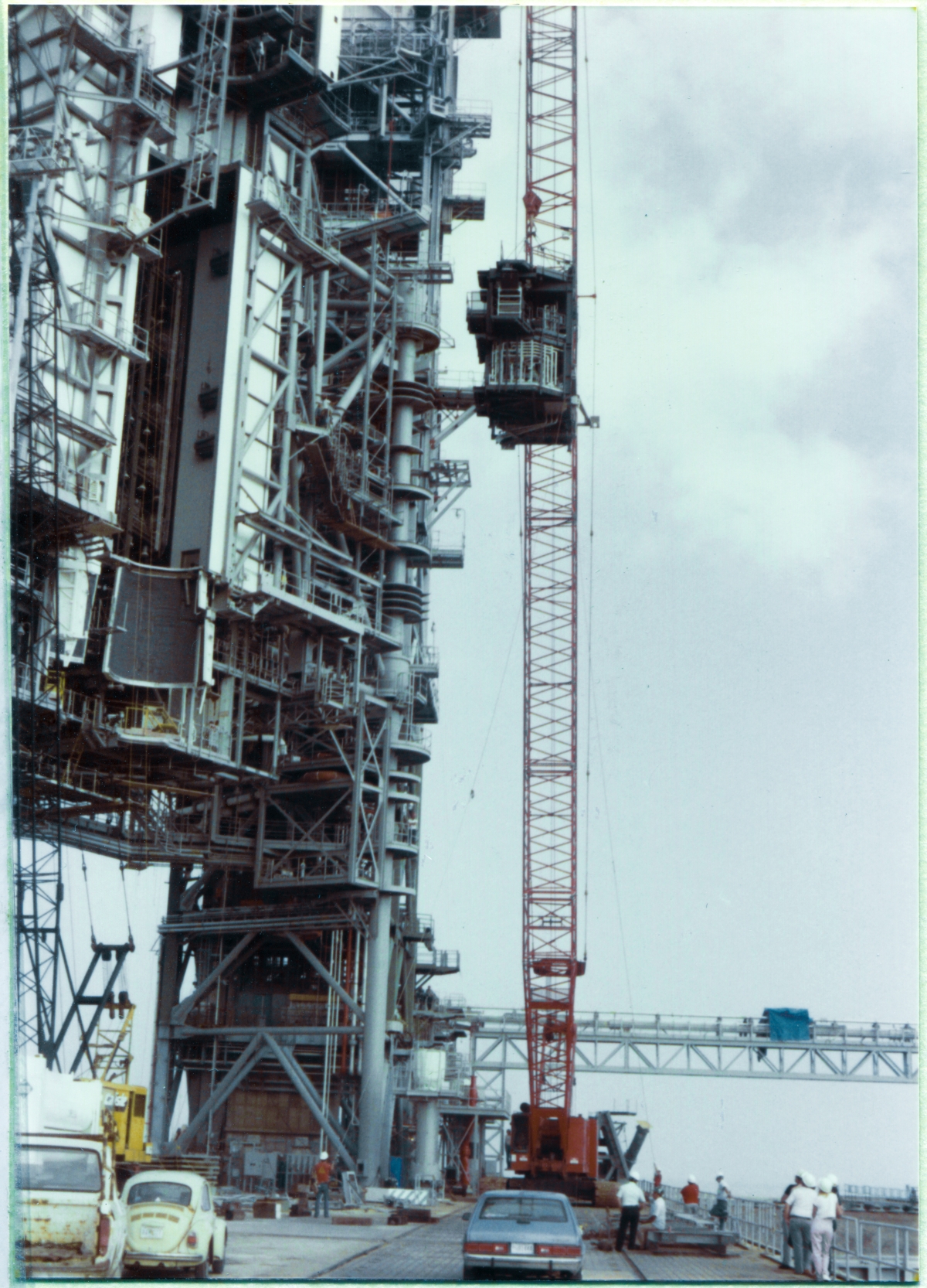 Image 092. At Space Shuttle Launch Complex 39-B, Kennedy Space Center, Florida, the Orbiter Mid-Body Umbilical Unit Lift has proceeded to the point where the OMBUU has reached its final elevation, and will soon be swung in toward the Rotating Service Structure, with the crane operator booming right, to do so. The bottom of the OMBUU is now over 100 feet above the concrete of the Pad Deck, and a crew of Union Ironworkers working for Ivey Steel stands on the OMBUU Access Catwalk at Elevation 163'-9” on the face of the RSS, ready to proceed with the next phases of connecting it to the tower. On the ground, various individuals, representing differing contractor and government entities, continue to monitor the progress of the Lift. Photo by James MacLaren.